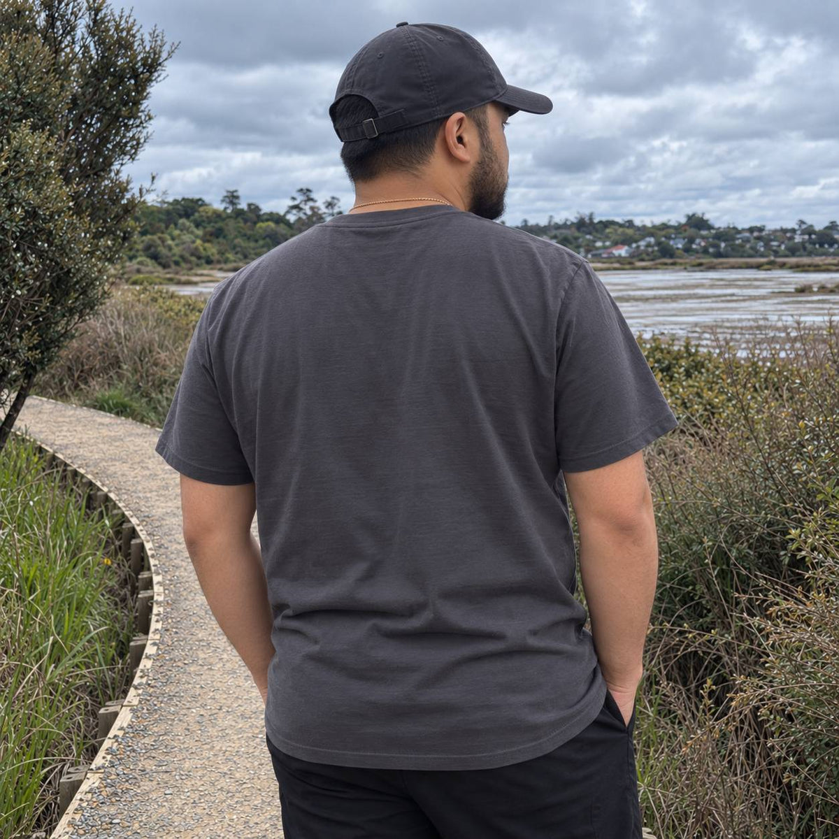 A man viewed from behind wearing a dark charcoal gray t-shirt and a black baseball cap, standing on a gravel path in an outdoor marsh landscape under a cloudy sky.