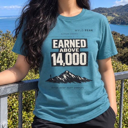 A woman wearing a teal graphic t-shirt with a mountain design and the text 'EARNED ABOVE 14,000' and 'WYLD PEAK', photographed outdoors with a scenic lake and hills in the background.