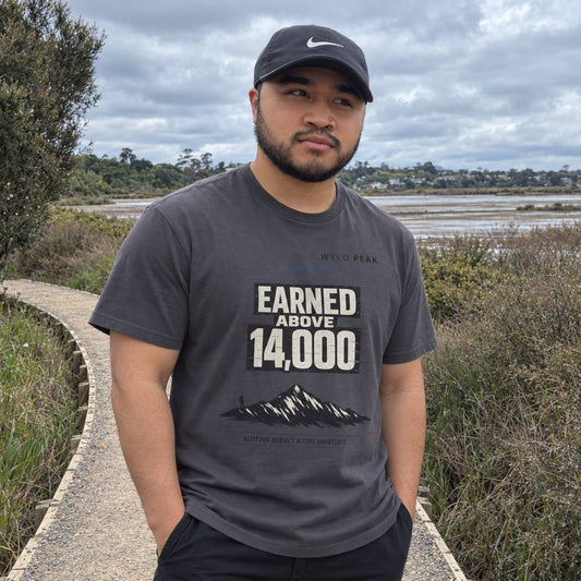 A man wearing a dark gray graphic t-shirt with 'EARNED ABOVE 14,000' and a mountain graphic, along with a black Nike cap, standing outdoors on a path with marshlands in the background.