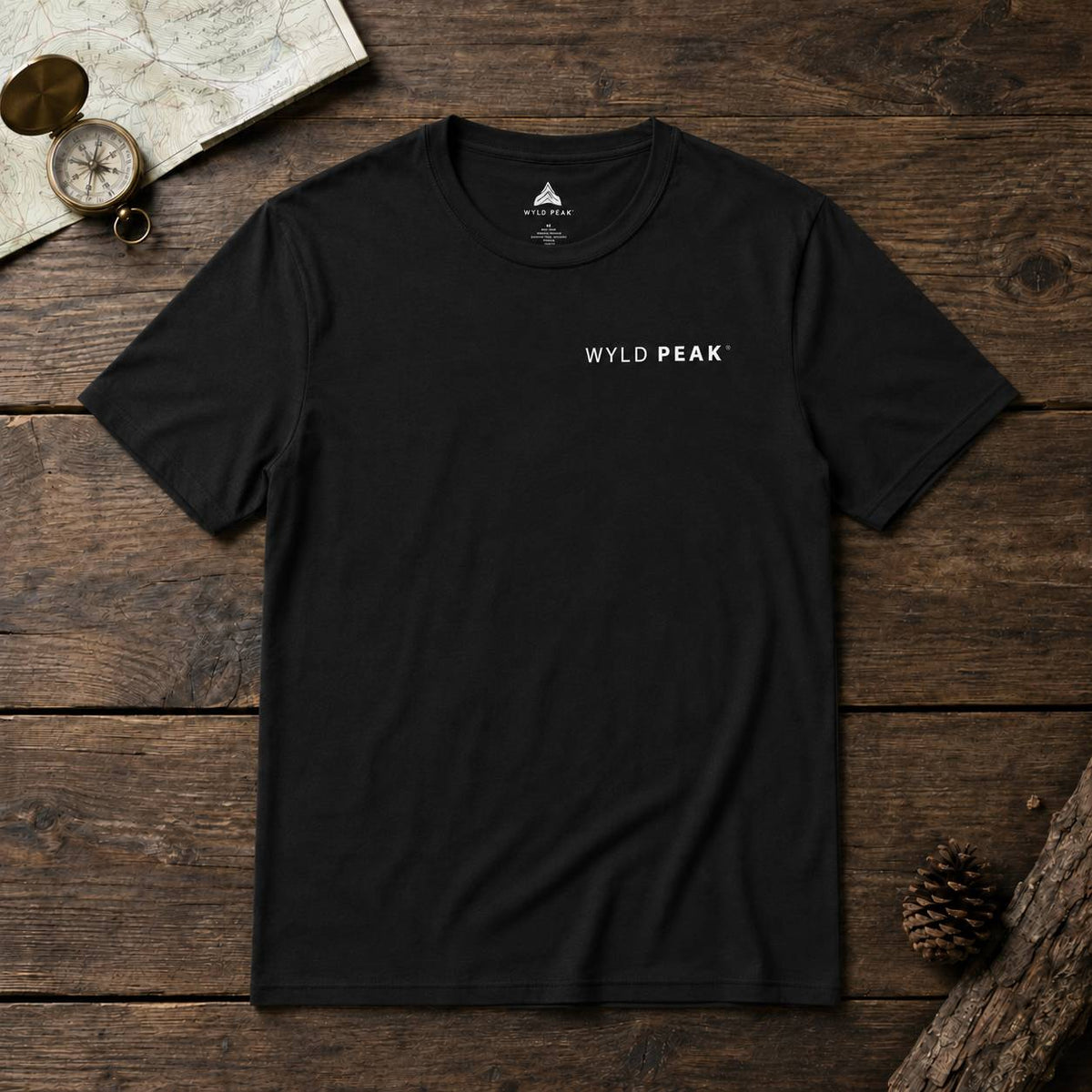 A black short-sleeve crewneck t-shirt with 'WYLD PEAK' printed in white on the chest, laid flat on a rustic dark wood background with a map, compass, and pinecone props.