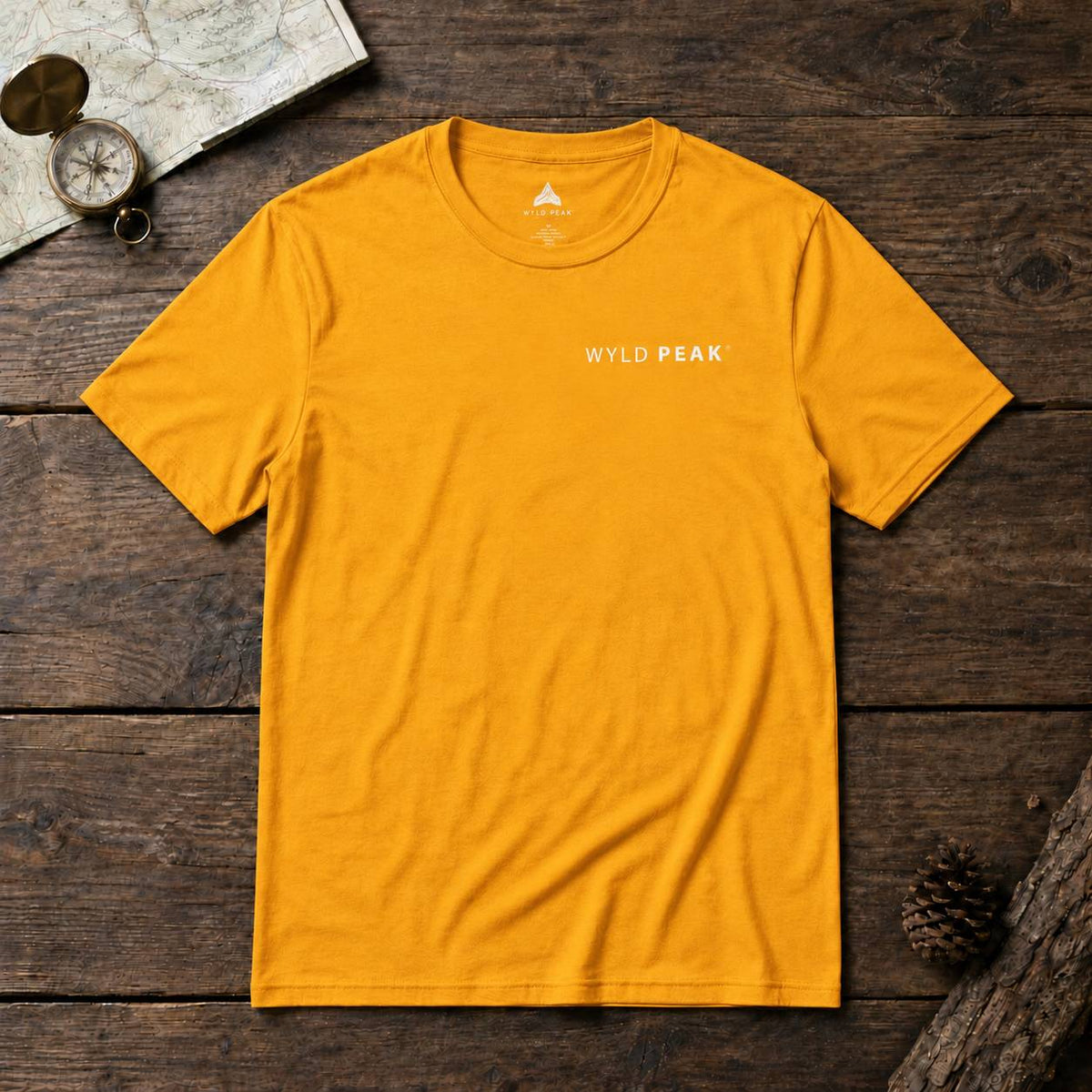 A mustard yellow crewneck t-shirt with 'WYLD PEAK' printed in white on the chest, laid flat on a dark rustic wooden background with a map, compass, and pinecone props.