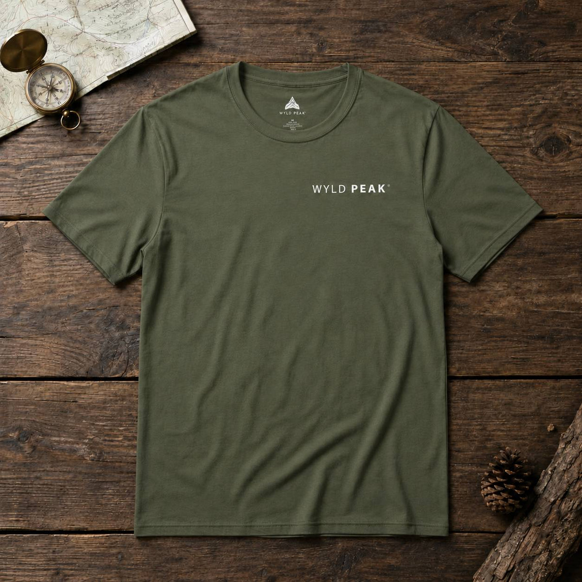 An olive green crewneck t-shirt with 'WYLD PEAK' printed in white on the left chest, laid flat on a rustic dark wood background with a map, compass, and pinecone props.