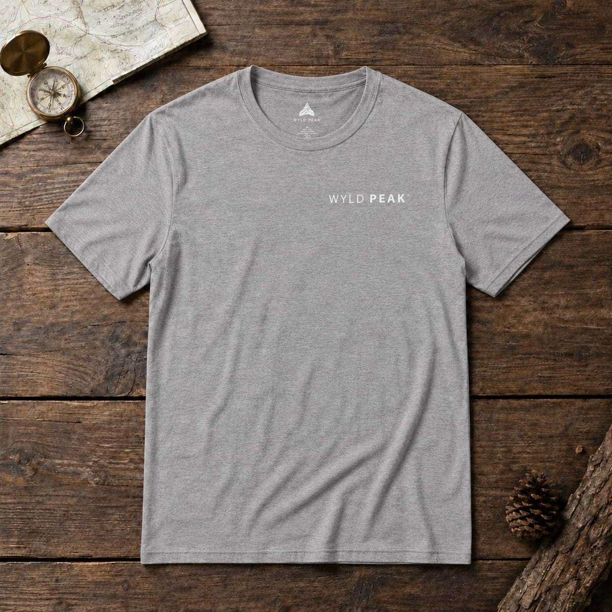 A heather gray crewneck t-shirt with 'WYLD PEAK' printed in white on the left chest, laid flat on a rustic dark wood background with a map, compass, and pinecone props.