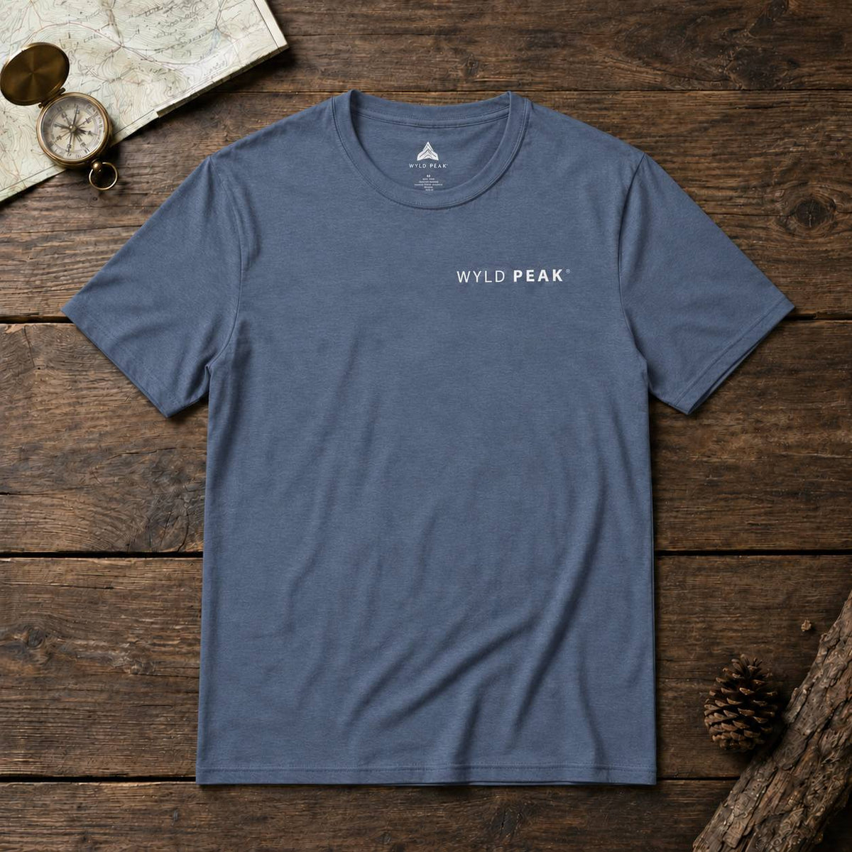 A slate blue crewneck t-shirt with 'WYLD PEAK' printed in white on the chest, laid flat on a rustic dark wood background with a map, compass, and pinecone props.