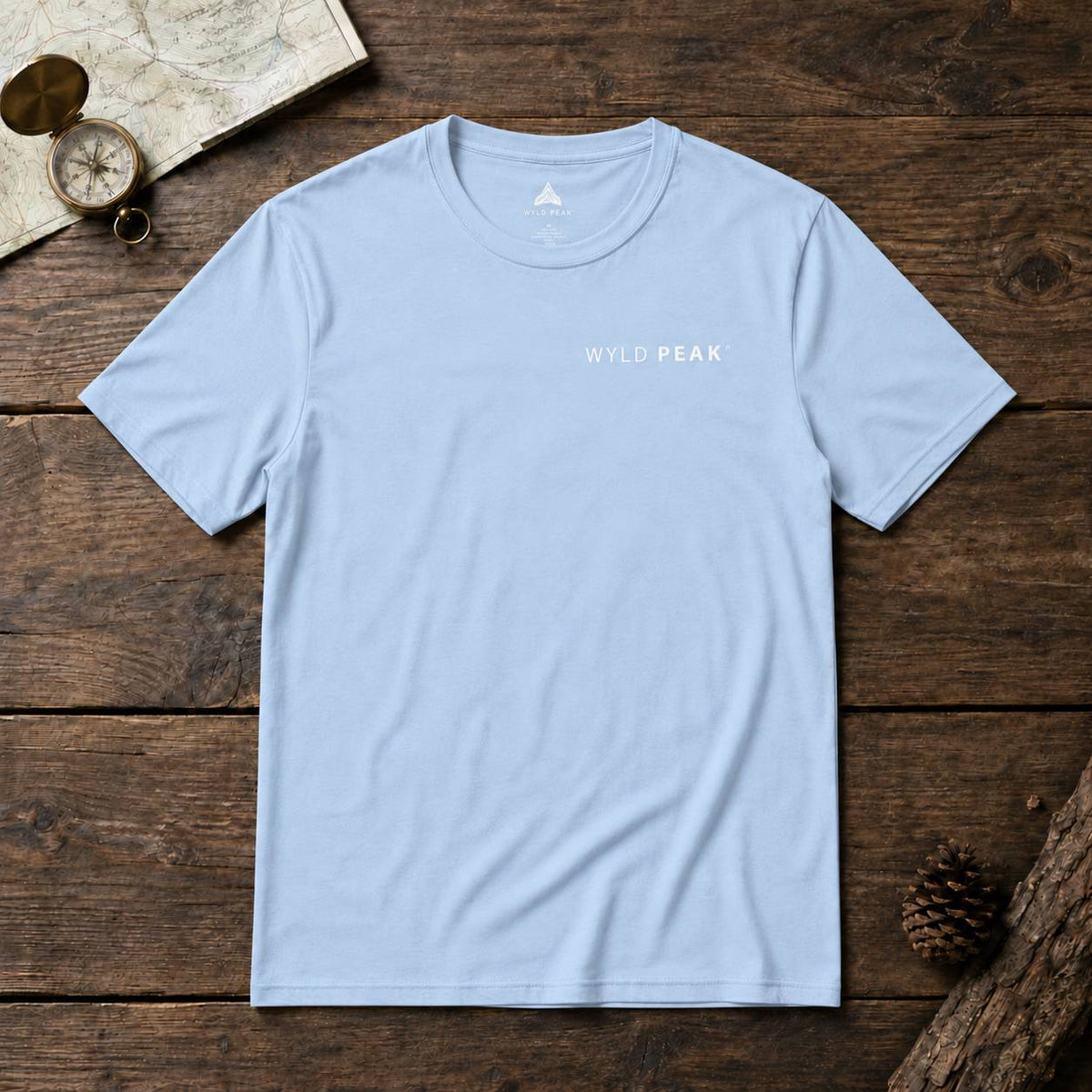 A light blue crewneck t-shirt with 'WYLD PEAK' printed in white on the chest, laid flat on a rustic dark wood background with a map, compass, and pinecone props.