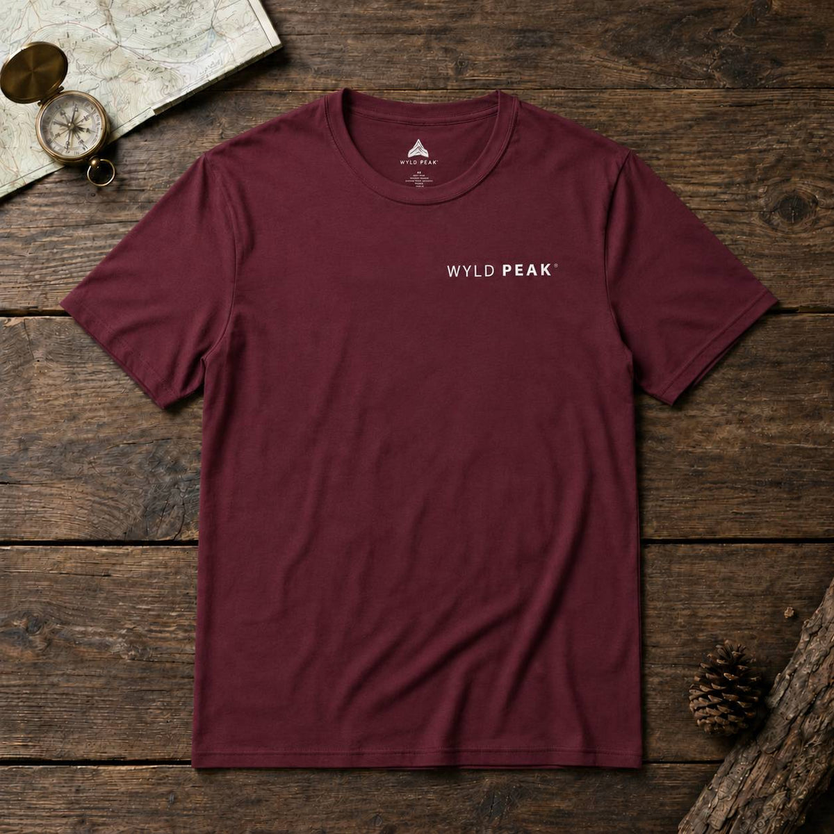 A maroon short-sleeve crewneck t-shirt with 'WYLD PEAK' printed in white on the left chest, laid flat on a rustic wooden background with a map, compass, and pinecone props.