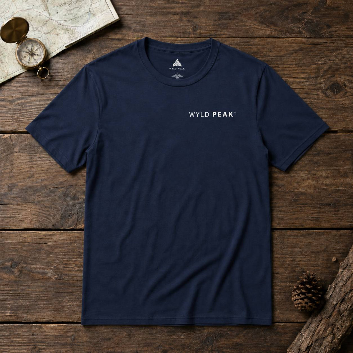 A navy blue crewneck t-shirt with 'WYLD PEAK' printed in white on the left chest, laid flat on a rustic wooden surface with a map, compass, and pinecone props.
