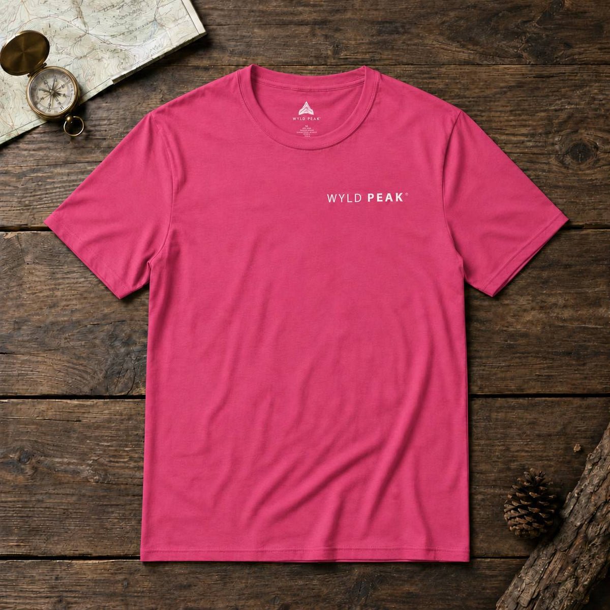 A bright pink crewneck t-shirt with 'WYLD PEAK' printed in white on the left chest, laid flat on a rustic dark wood background with a map, compass, and pinecone props.