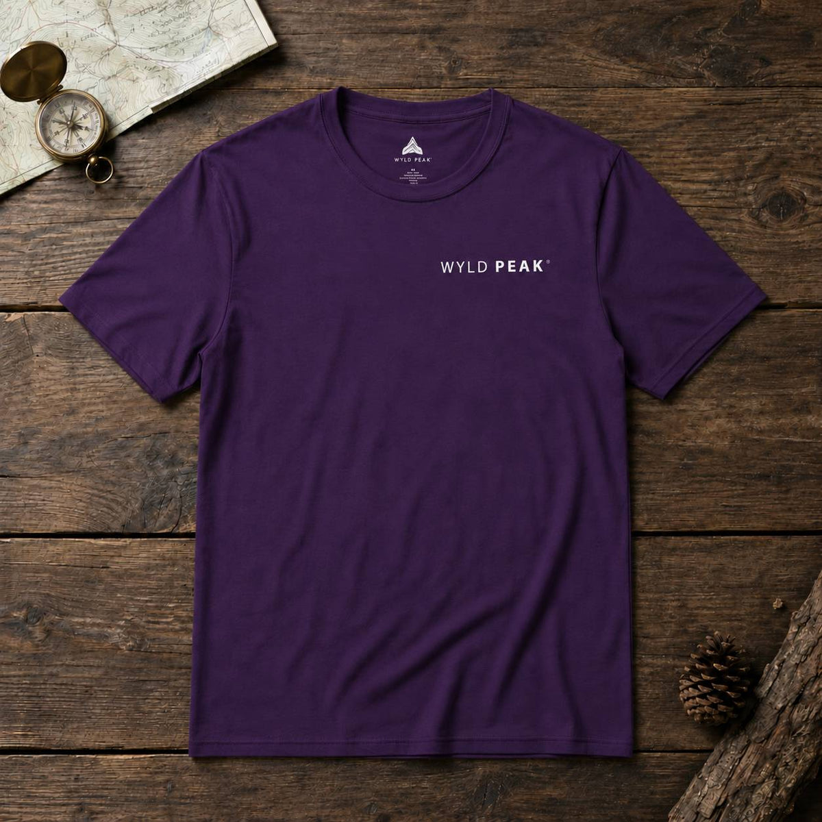 A purple crewneck t-shirt with 'WYLD PEAK' printed in white on the chest, laid flat on a rustic wooden surface with vintage map, compass, and pinecone props.