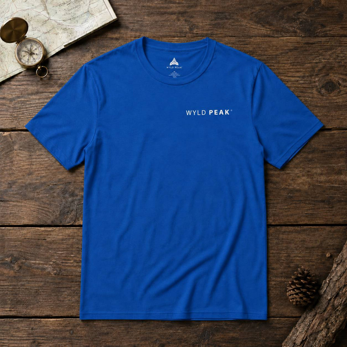 A royal blue crewneck t-shirt with 'WYLD PEAK' printed in white on the chest, laid flat on a rustic wooden surface with a vintage compass, map, and pinecone props.