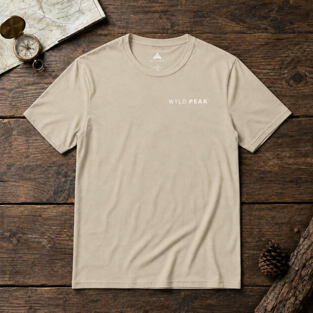 A beige crewneck t-shirt with 'WYLD PEAK' printed in white on the chest, laid flat on a rustic dark wood background with a map, compass, and pinecone props.