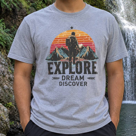A man wearing a heather gray t-shirt with a retro-style graphic of a hiker in front of mountains and a sunset, with the text 'EXPLORE. DREAM.