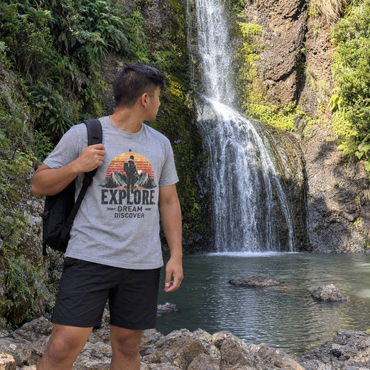 A man wearing a gray graphic t-shirt and black shorts stands on rocks in front of a waterfall, carrying a black backpack. The t-shirt features a colorful sunset graphic with the words 'EXPLORE DREAM DISCOVER'.