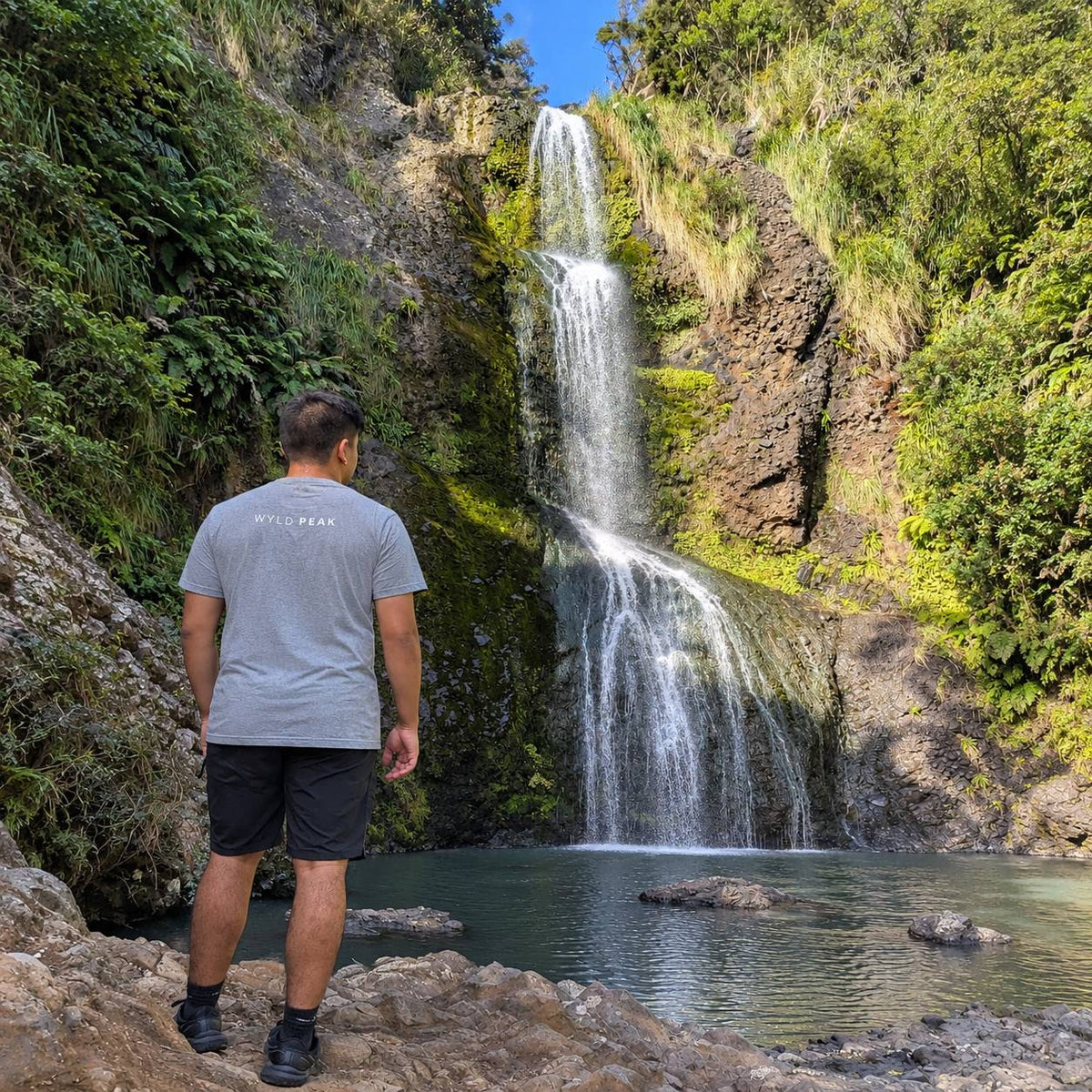 A man wearing a gray t-shirt and black shorts stands on a rocky ledge, looking at a tall waterfall cascading into a pool in a lush green outdoor setting.