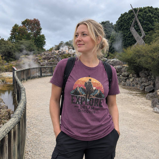 A woman wearing a heathered purple graphic t-shirt with a sunset mountain design and the words 'EXPLORE DREAM DISCOVER' stands on a gravel path outdoors.