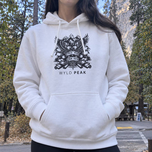 A person wearing a white pullover hoodie with a black graphic print on the chest that reads 'WYLD PEAK' and features an eagle, mountain, and crossed rifles design. The photo is taken outdoors in a forested area with trees and a mountain visible in the background.