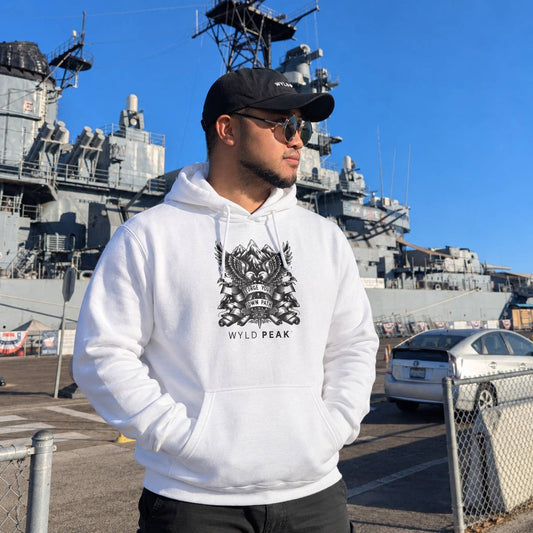 A man wearing a white pullover hoodie with a black graphic print and the text 'WYLD PEAK' stands outdoors in front of a large gray naval ship under a clear blue sky.