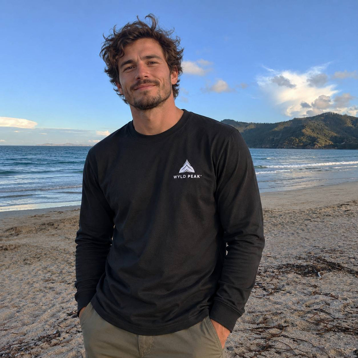 A man standing on a sandy beach wearing a black long-sleeve t-shirt with a white Wyld Peak logo on the left chest, with the ocean and mountains in the background.