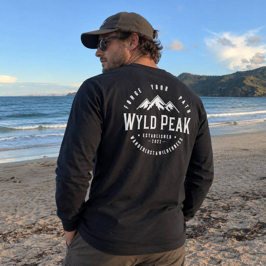 A man standing on a sandy beach with the ocean and mountains in the background, wearing a black long-sleeve shirt with a large white 'WYLD PEAK' mountain graphic on the back, a brown cap, and sunglasses.