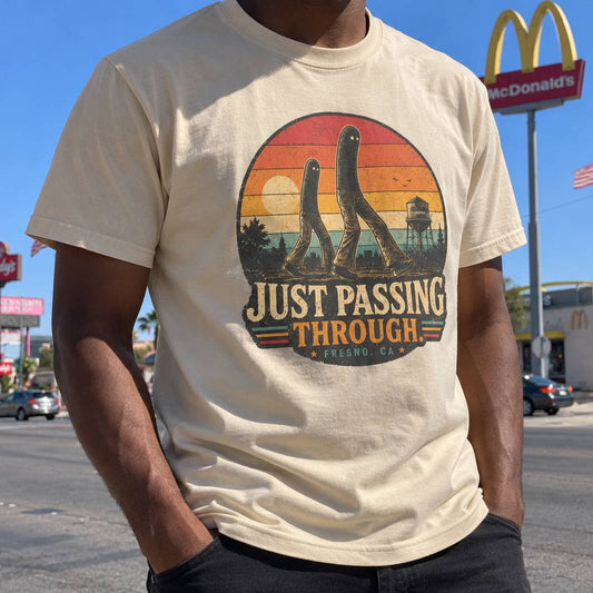 A man wearing a cream-colored graphic t-shirt with a retro-style print of two walking figures in a sunset circle and the text 'JUST PASSING THROUGH' and 'FRESNO, CA'. The background shows a street scene with a McDonald's sign visible.