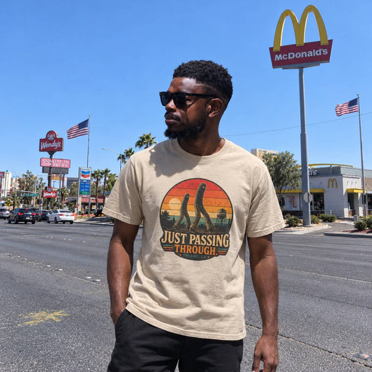 A man wearing a beige graphic t-shirt with a 'Just Passing Through' Nightcrawler print, standing on a sunny street in front of a McDonald's sign.