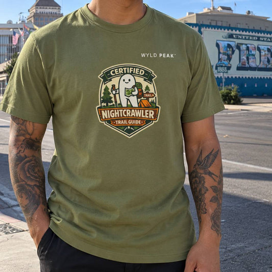 A man wearing an olive green t-shirt with a 'Certified Nightcrawler Trail Guide' graphic on the chest, standing outdoors on a street with a blue building in the background.