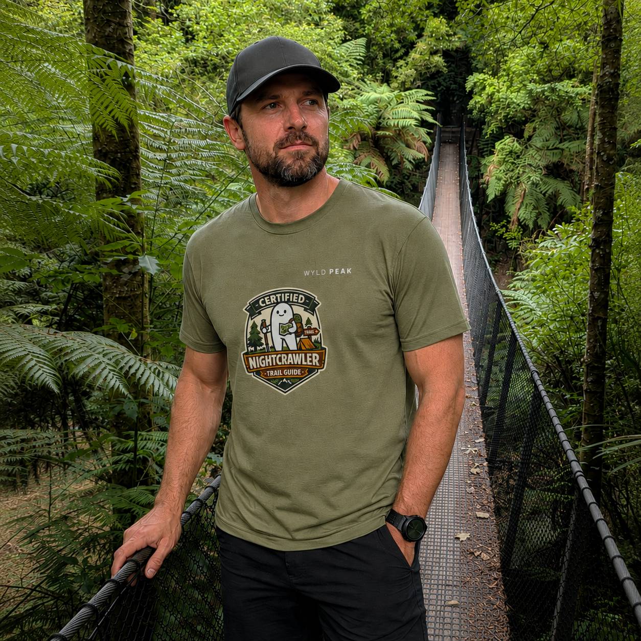 A man wearing an olive green t-shirt with a 'Fresno Nightcrawler' graphic, standing on a suspension bridge in a lush green forest.