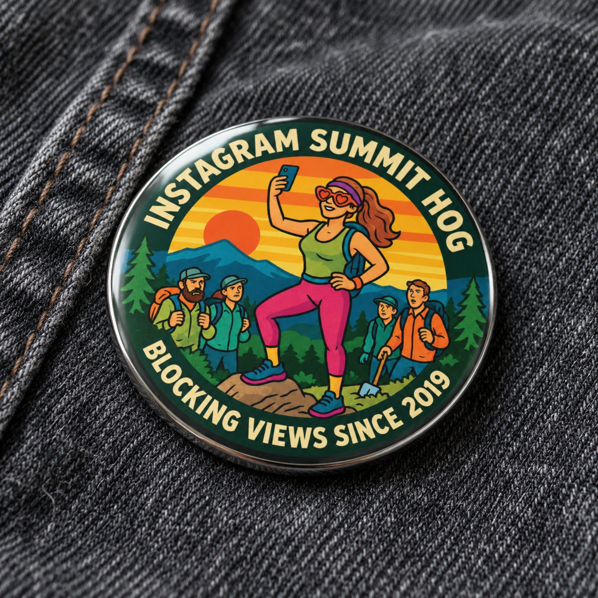 A round enamel pin with a colorful hiking illustration and text reading 'Summit Hog Blocking Views Since 2019,' photographed resting on dark gray denim fabric.