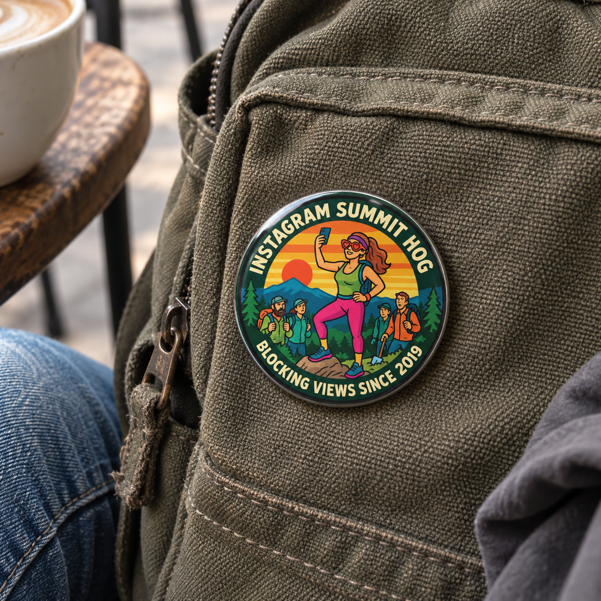 A circular enamel pin featuring a colorful illustration of a woman taking a selfie on a mountain with the text 'SUMMIT HOG' and 'BLOCKING VIEWS SINCE 2019', attached to an olive green canvas backpack. A person's jeans and a coffee cup are partially visible in the lifestyle setting.
