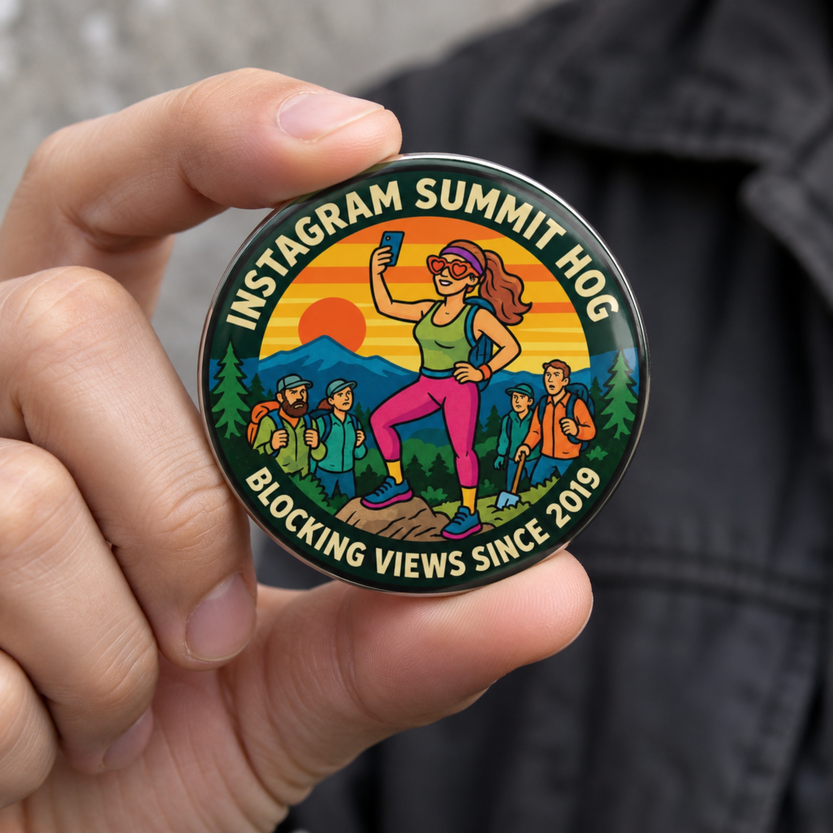 A hand holding a round enamel pin with a colorful illustration of a woman blocking a sunset and the text 'Summit HOG Blocking Views Since 2019'.