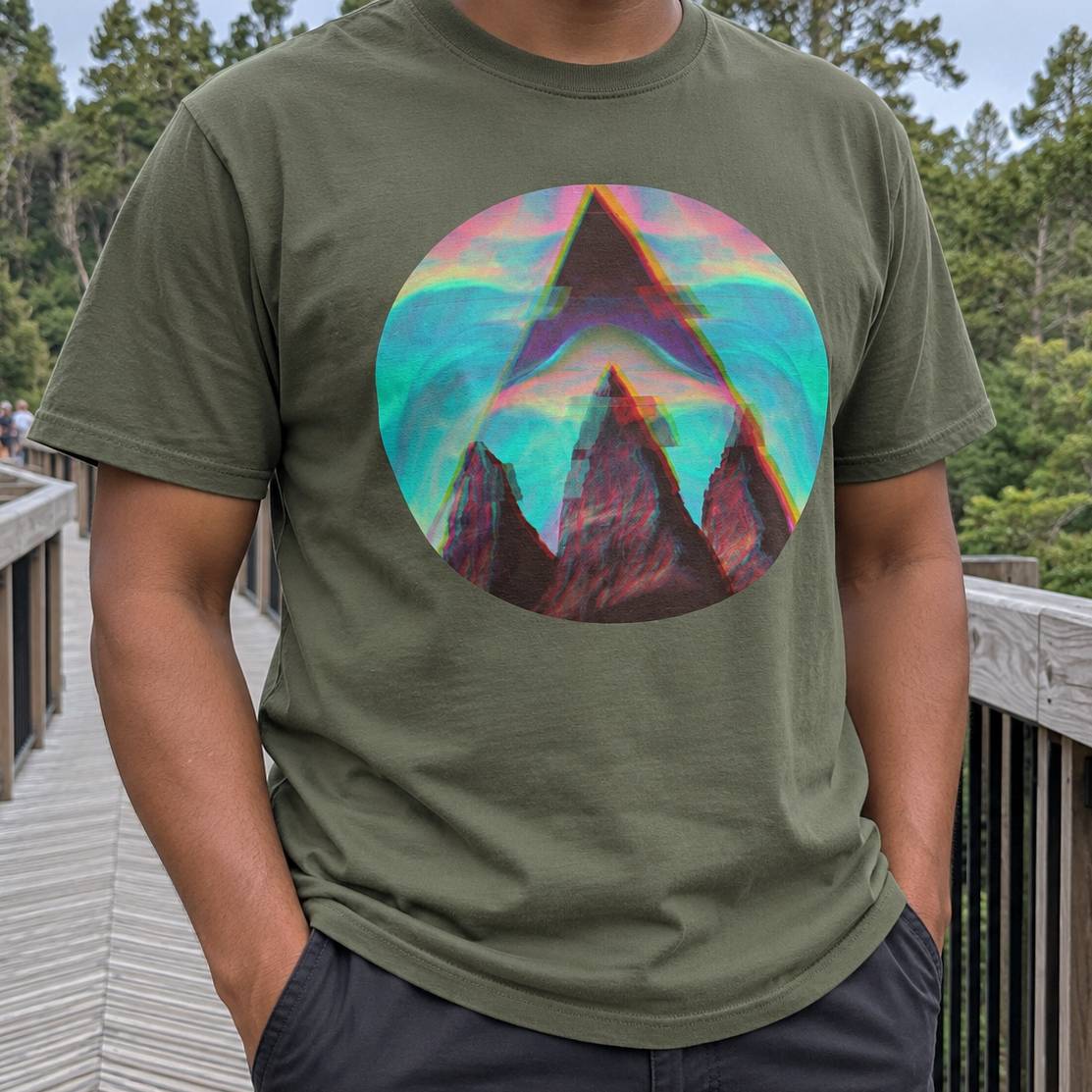 A person wearing an olive green t-shirt with a colorful circular graphic of a mountain peak under a rainbow gradient sky, standing on a wooden boardwalk with trees in the background.