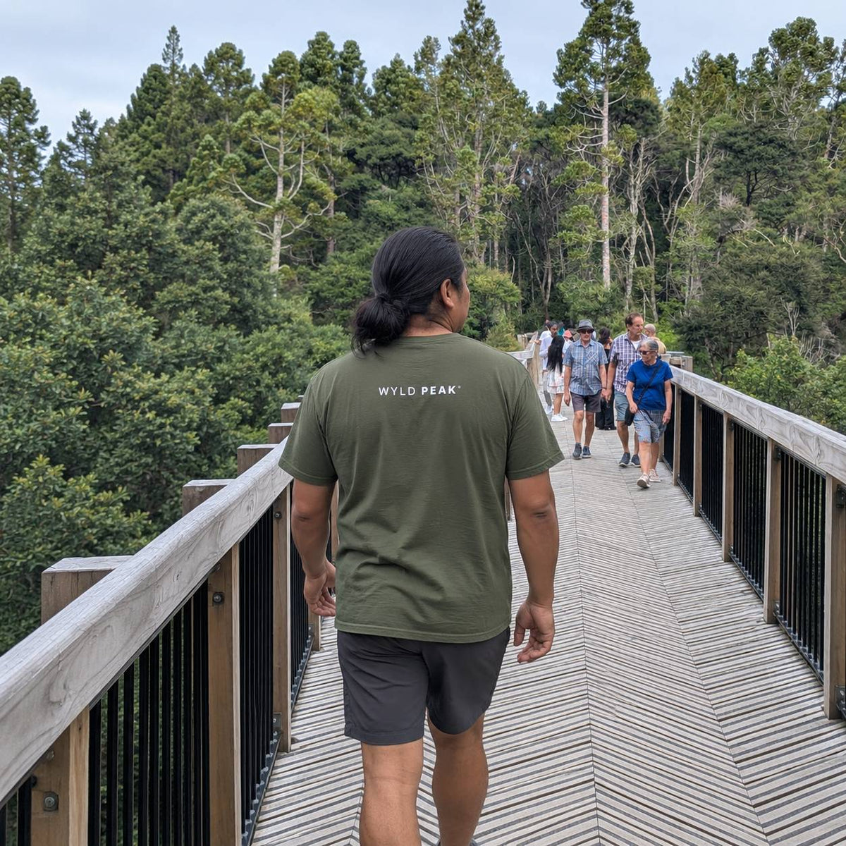 A man wearing an olive green t-shirt with 'WYLD PEAK' printed on the back walks along a wooden boardwalk surrounded by a dense green forest. Other people are visible walking in the background.