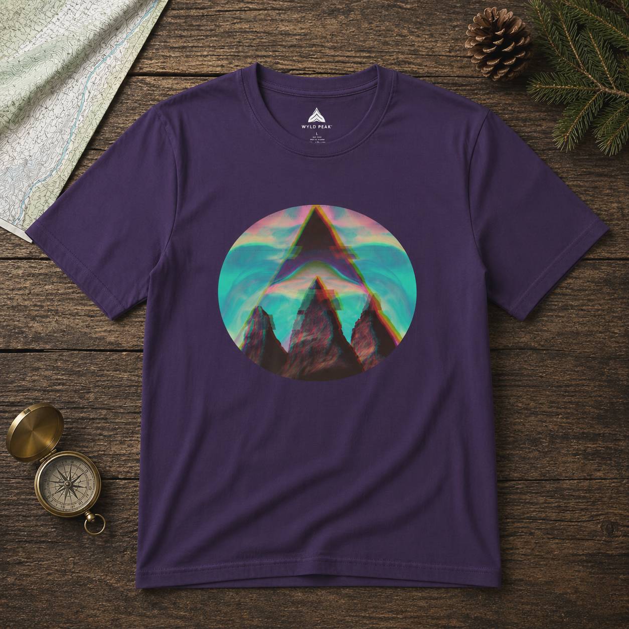 Purple t-shirt with a mountain graphic on a wooden surface with a compass and pine cone.