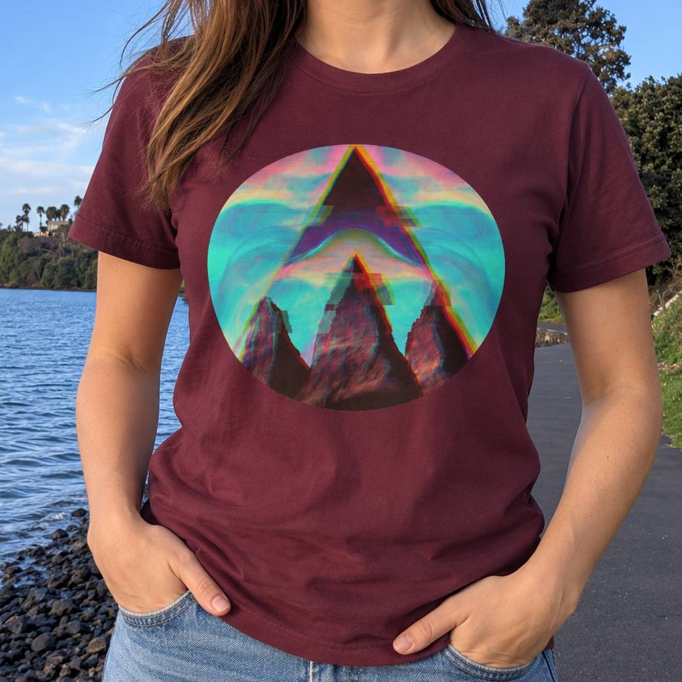 A woman wearing a maroon crewneck t-shirt with a colorful circular graphic of a stylized mountain peak in front of a teal and pink sky. The background shows a real outdoor environment with water, trees, and a path, making the subject type 'Lifestyle'.