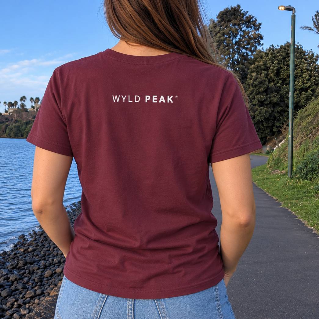 A woman viewed from behind wearing a maroon short-sleeve t-shirt with 'WYLD PEAK' printed in white across the upper back, standing outdoors next to a body of water and a paved path.