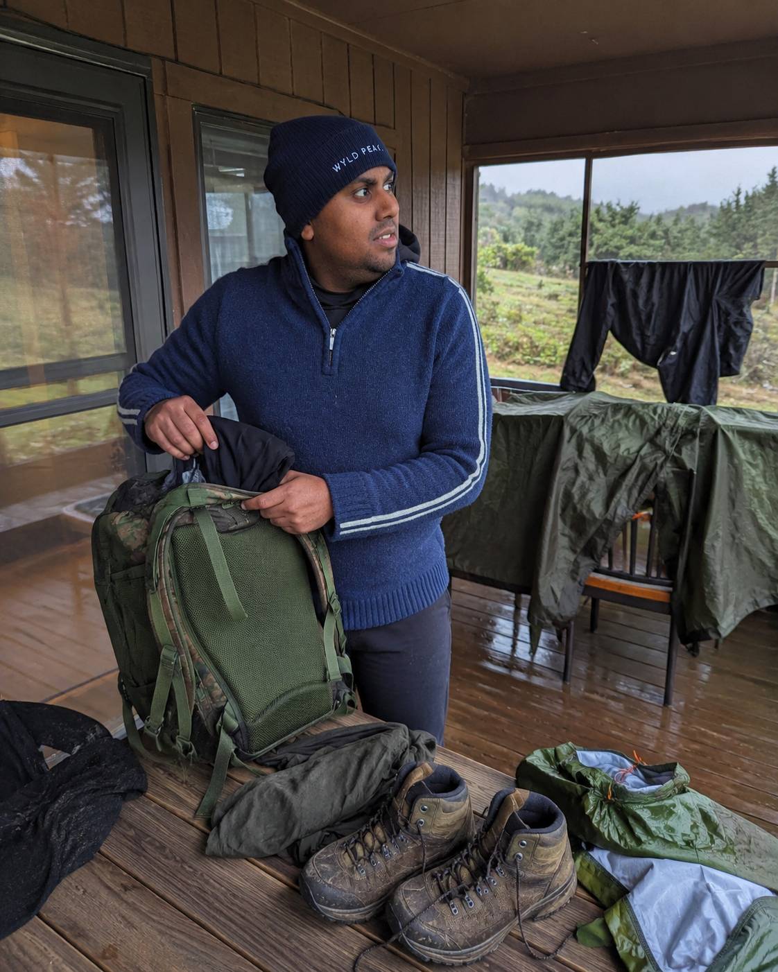 Person preparing for an outdoor adventure with backpack and boots inside a cabin.