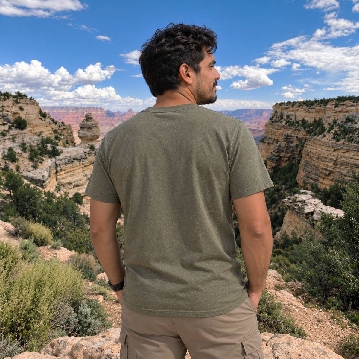 A man standing on a canyon cliffside, viewed from behind, wearing an olive green t-shirt and khaki cargo pants under a blue sky with white clouds.