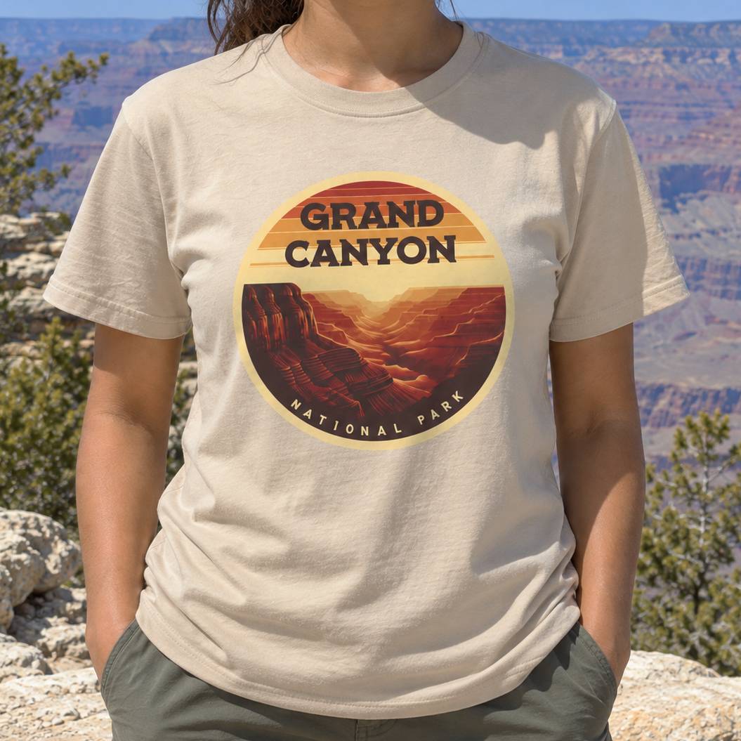 A person outdoors wearing a light beige T-shirt with a circular Grand Canyon National Park graphic on the chest, set against a canyon landscape background.