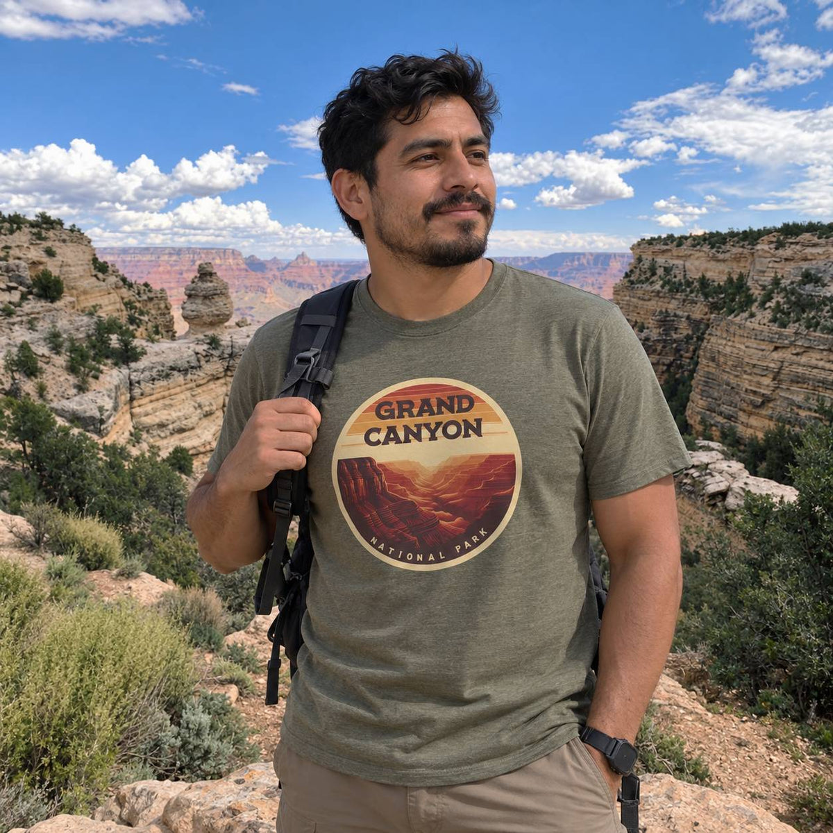 A man wearing an olive green t-shirt with a circular 'Grand Canyon National Park' graphic, standing outdoors in a scenic canyon landscape under a blue sky.