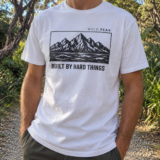 A man wearing a white crewneck t-shirt with a black graphic of mountains and the text 'WYLD PEAK' and 'BUILT BY HARD THINGS', photographed outdoors in a wooded setting.