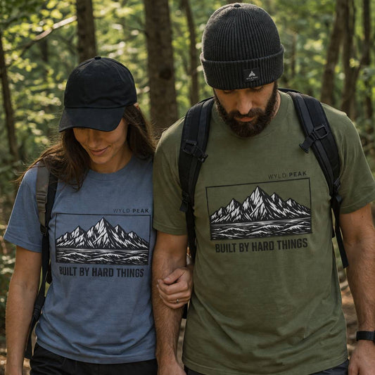 Two people hiking in a forest wearing Wyld Peak t-shirts with mountain designs.
