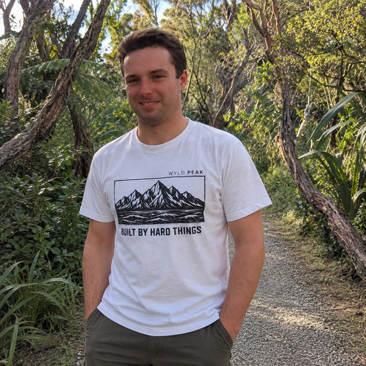 A man standing on a gravel path in a wooded outdoor setting, wearing a white crewneck t-shirt with a black graphic of mountains and the text 'WYLD PEAK' and 'BUILT BY HARD THINGS'.