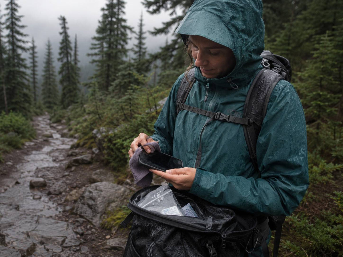 Person in a green raincoat with a backpack using a device on a forest path.