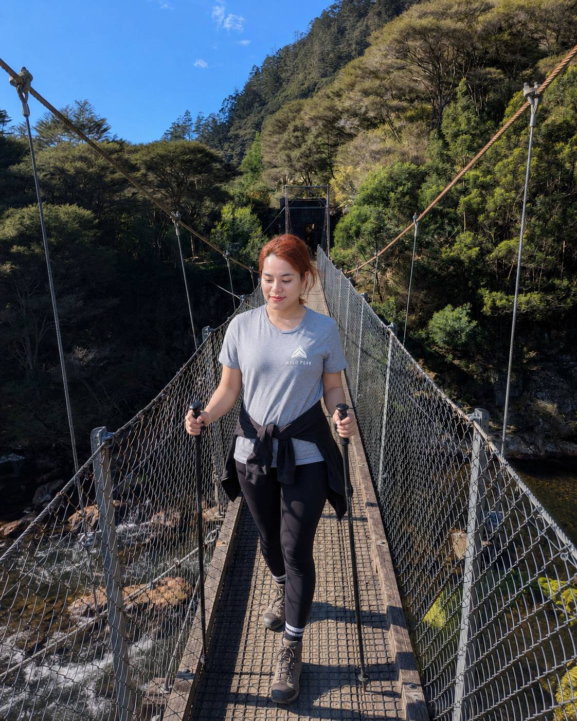 A woman hiking on a suspension bridge over a river, wearing a light gray t-shirt, black leggings, and carrying a black jacket around her waist, with a lush green mountain backdrop.