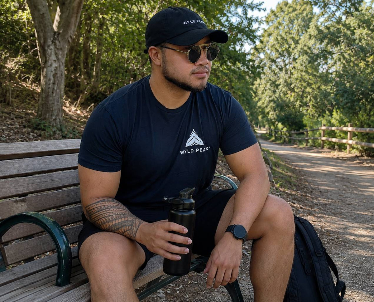 Man sitting on a bench outdoors holding a water bottle, wearing a black t-shirt with a logo and a cap.