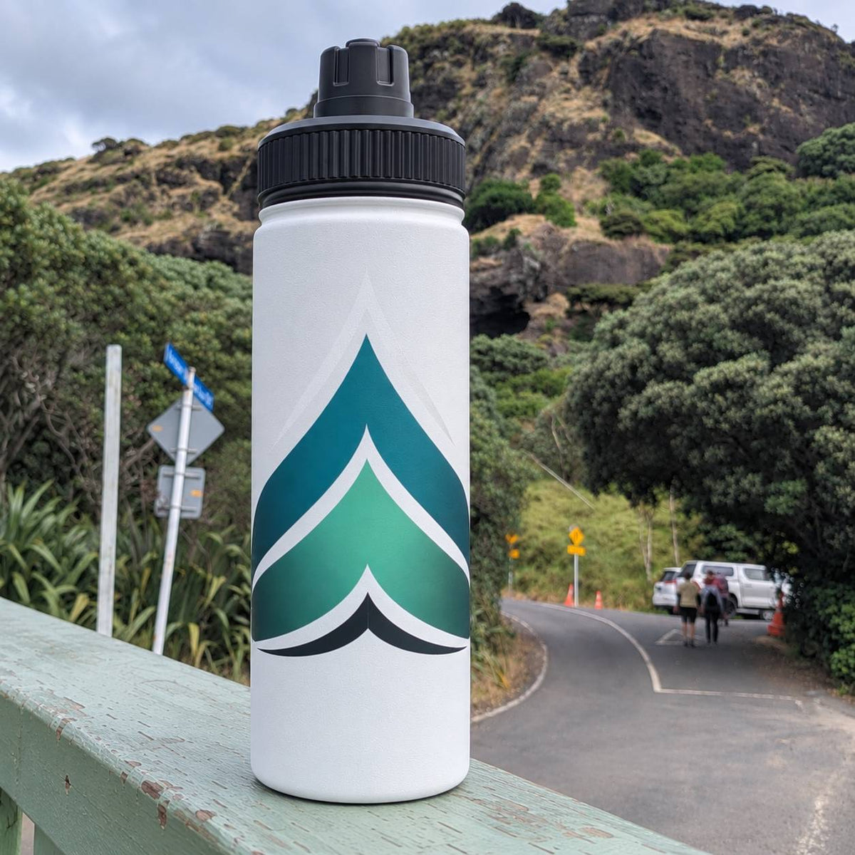 A white water bottle with a black cap stands on a weathered green wooden railing, set against a scenic outdoor background of a winding road, trees, and rocky hills.