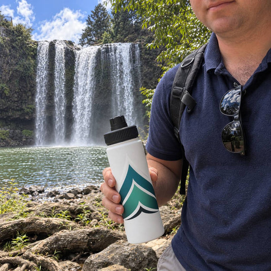 A man wearing a navy blue polo shirt and holding a white water bottle with a green and black mountain logo, standing outdoors in front of a waterfall and trees.