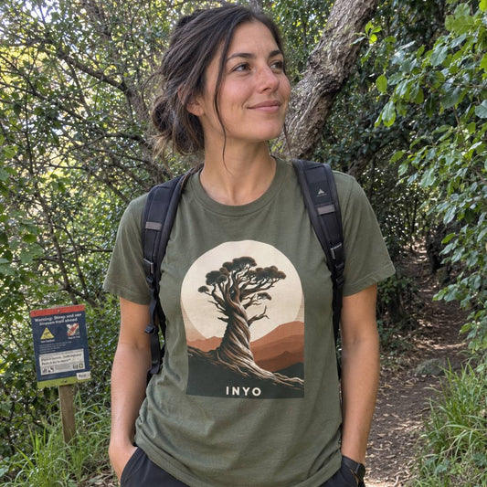 A woman wearing an olive green t-shirt with a circular graphic of a gnarled tree and the word 'INYO' on the front, standing on a wooded trail with a backpack.