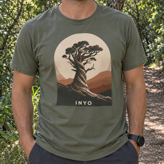 A man wearing an olive green t-shirt with a vintage-style graphic of a gnarled tree in a landscape with the text 'INYO' below it, set against a forest background.