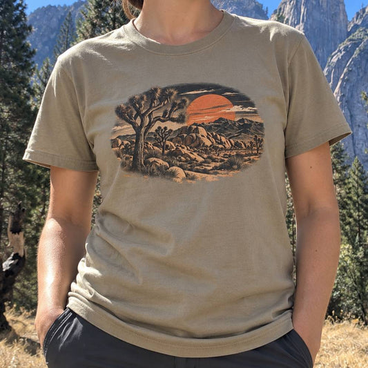 A person wearing a beige graphic t-shirt with a desert sunset print, photographed outdoors in a mountain and pine forest setting.